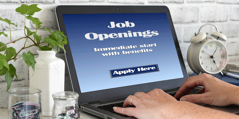 A laptop open to the words "Job Openings. Immediate start with benefits. Apply here." The desk has a white clock, fake plant, and paperclips and pins on it.